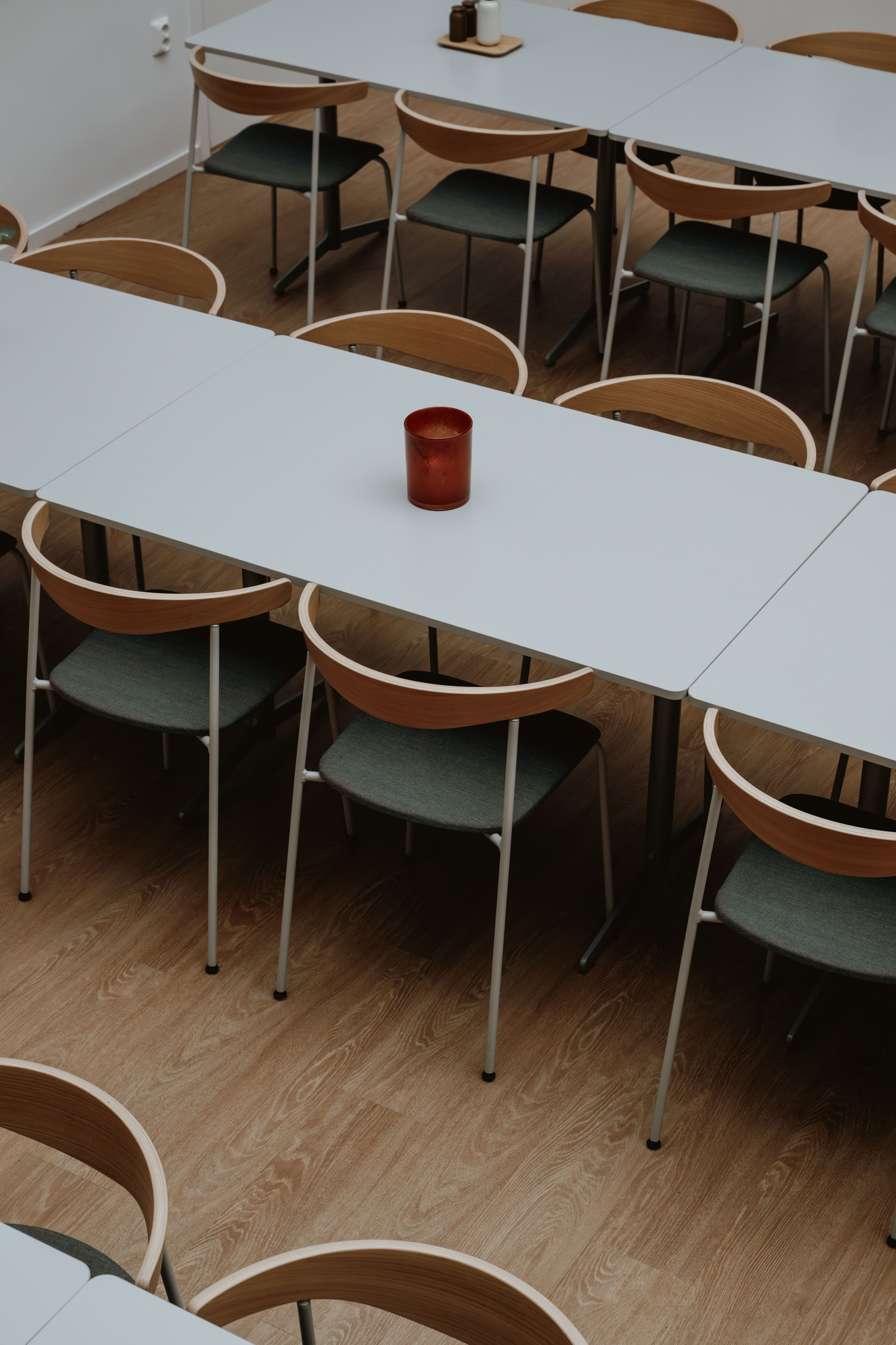 Overhead view of a cafeteria with light grey tables, chairs with wooden backs and grey seats, a red candle holder, and a wooden floor.