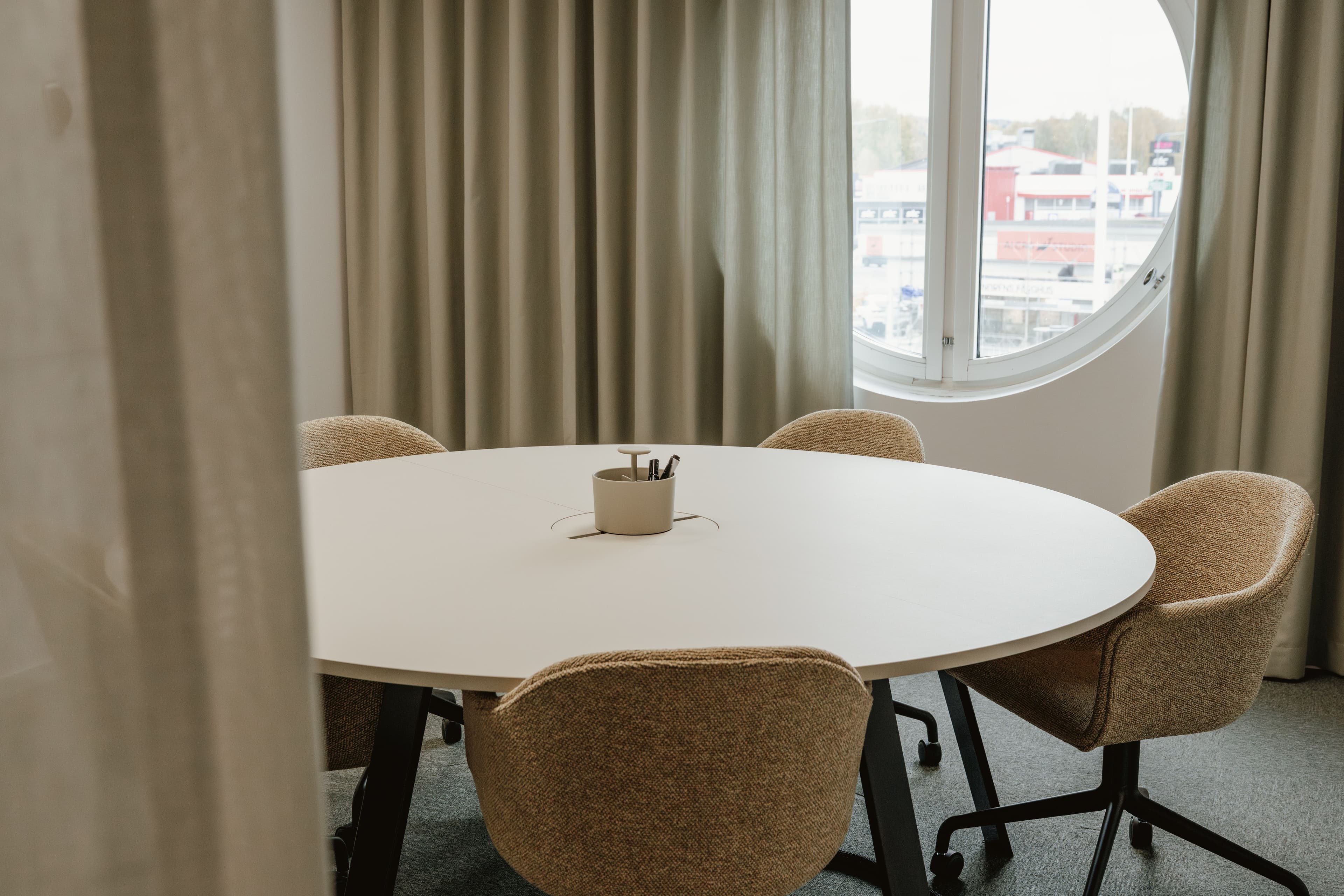 A modern meeting room with a round white table, four beige chairs, and a large circular window.